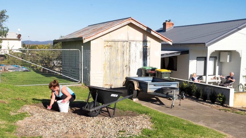 A woman digging along the verge on a suburban street, in the background a couple eat breakfast on the verandah.