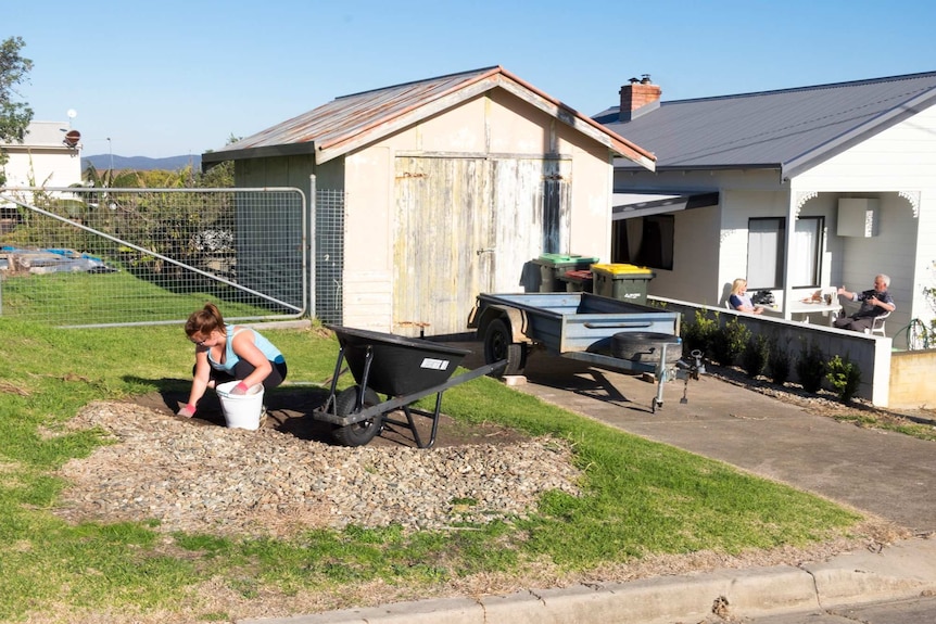 A woman digging along the verge on a suburban street, in the background a couple eat breakfast on the verandah.