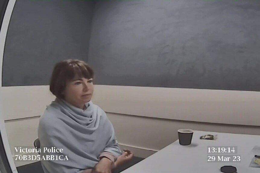A woman with brown hair wearing light blue sits in a police interview room.