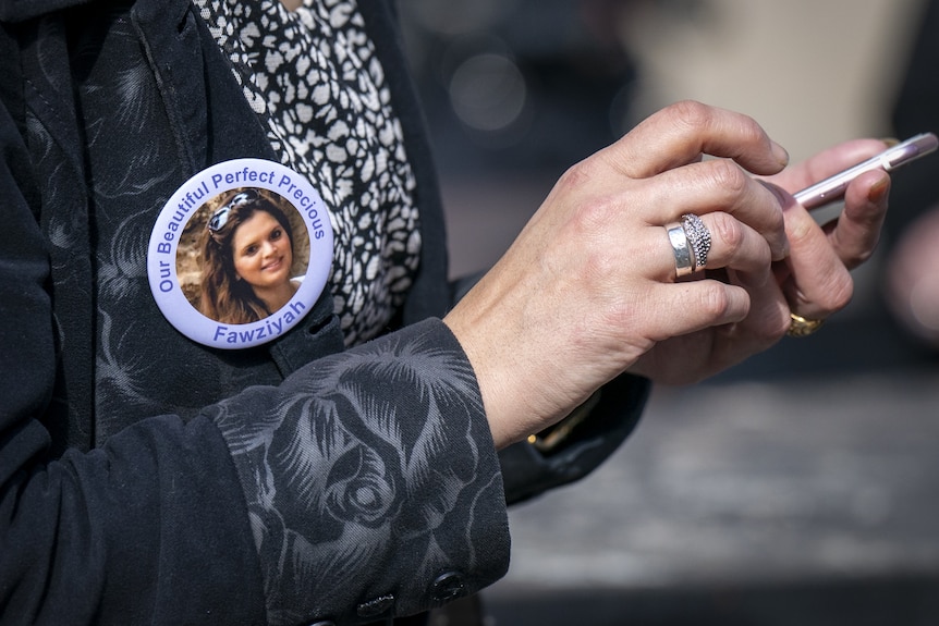 A person wearing a badge that reads "Our beautiful perfect precious Fawziyah", while also holding a phone.