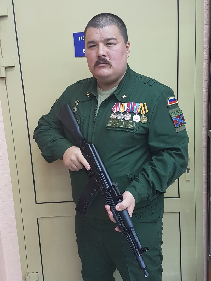 Man with shaved head and moustache wears green military outfit, holding machine gun, standing in front of door.
