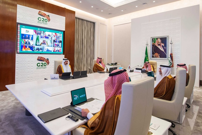 Saudi King Salman gives his opening remarks via video as he sits at large tables with a few other men.