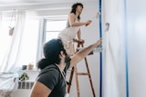 A couple uses rollers to paint a wall inside a house