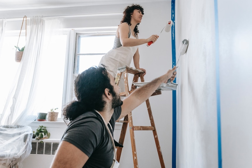 A couple uses rollers to paint a wall inside a house