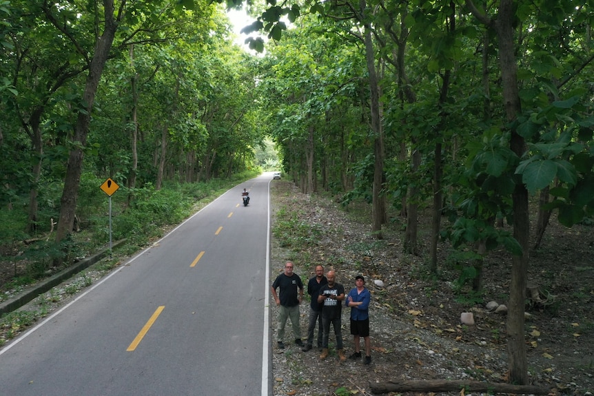 Four men stand on the side of the road surrounded by lush jungle. A motorbike is seen in the background.