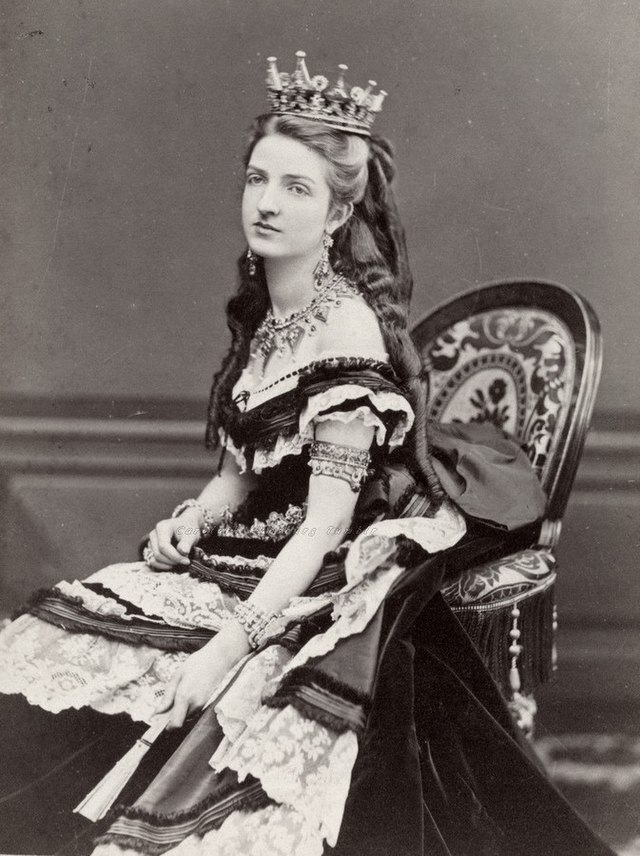 Black and white picture of Margherita of Savoy, Queen of Italy, sitting on a chair wearing a crown and ruffled dress
