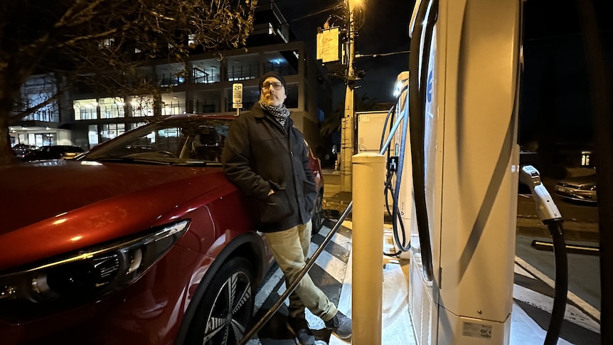 Michael Bond stands next to his red car at night at a charging station.