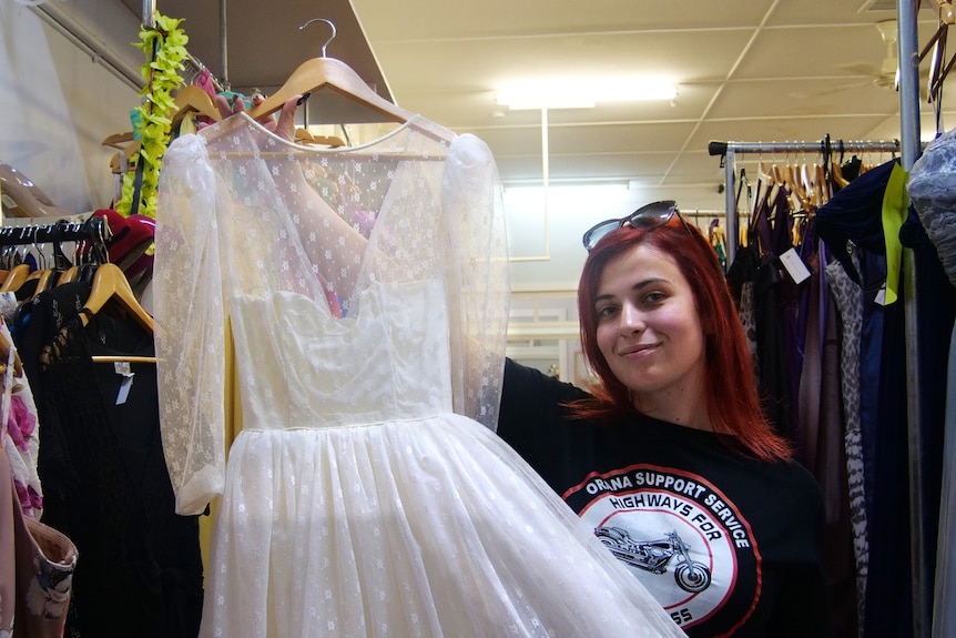A woman with red hair in a black t-shirt holds up a lacy cream wedding dress.