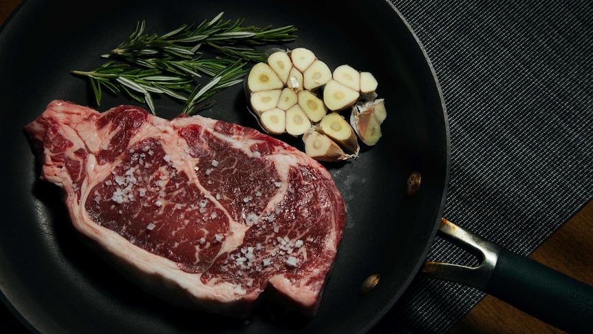 A raw steak in a frying pan, next to some rosemary and half a head of garlic