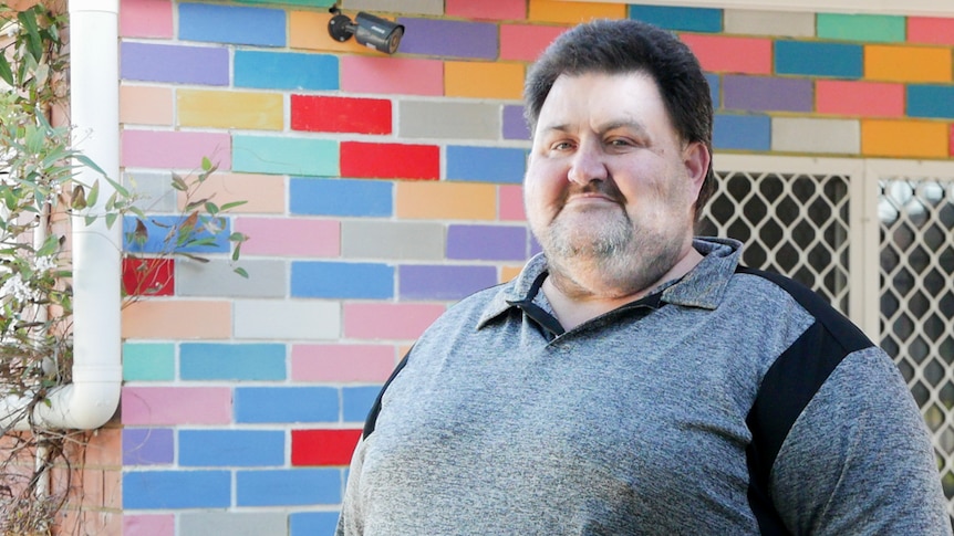 A man in a grey shirt standing in front of a colourful wall.