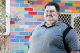 A man in a grey shirt standing in front of a colourful wall.