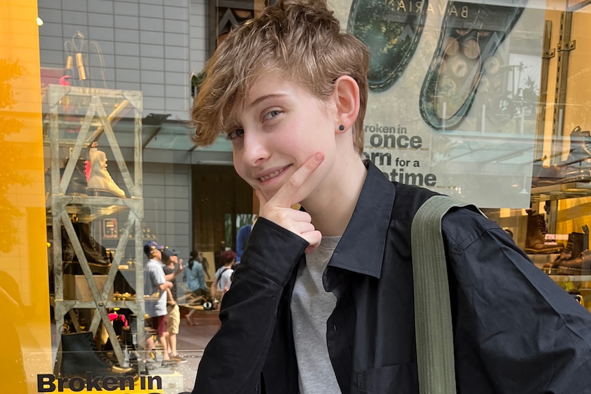 A smiling teenage boy holds his hand to his chin in a thinking gesture while standing outdoors in front of a shop.