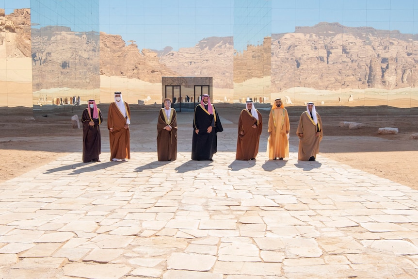 A group of robed Arab men stand in front of a giant mural of a sandy landscape