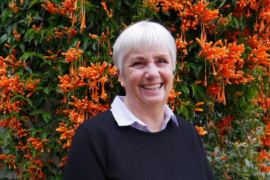 A woman in a black jumper with short white hair stands in front of orange flowers.