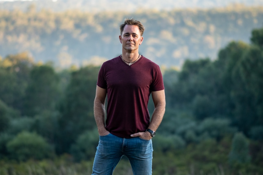 Wide shot of Jarrod standing on a lush hill, with valley of green trees lower behind him, wearing a red tshirt, hands in pockets