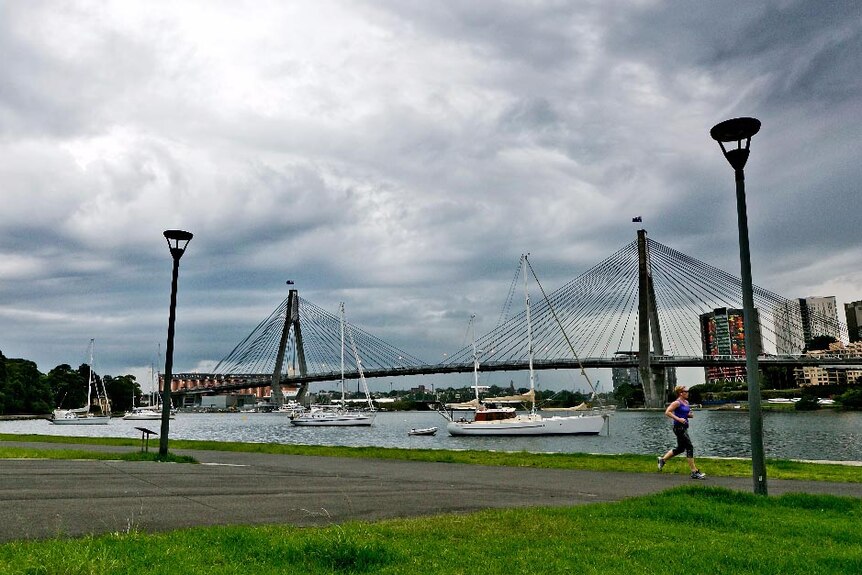 Storms roll in over the Anzac Bridge as a woman jogs along the waterfront in the foreground.