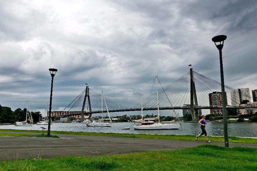 Storms roll in over the Anzac Bridge as a woman jogs along the waterfront in the foreground.