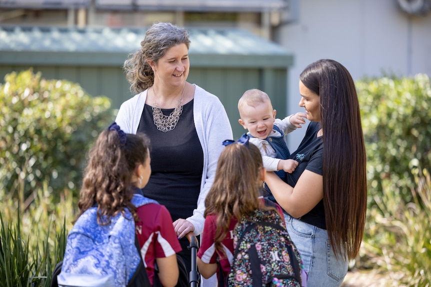 A teacher stands outside with two primary students and a mother holding a baby.