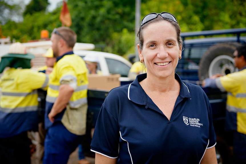 A smiling woman wearing a Charles Darwin University polo-shirt, with some men in high-vis in the background.