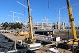 Cranes sit on a rail station construction site with Perth Stadium in the distance under a blue sky.