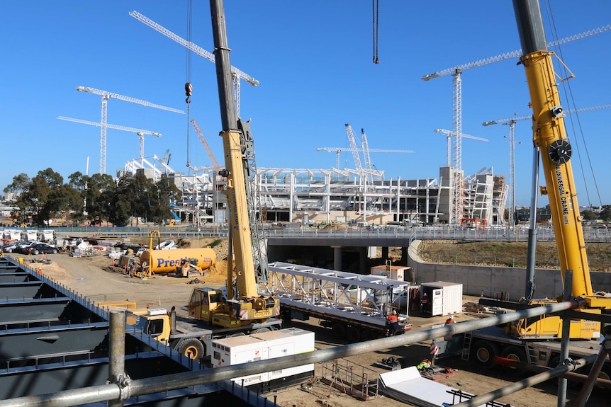 Cranes sit on a rail station construction site with Perth Stadium in the distance under a blue sky.