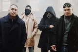 Four men in dark clothes, two in hoods, standing inside a church.