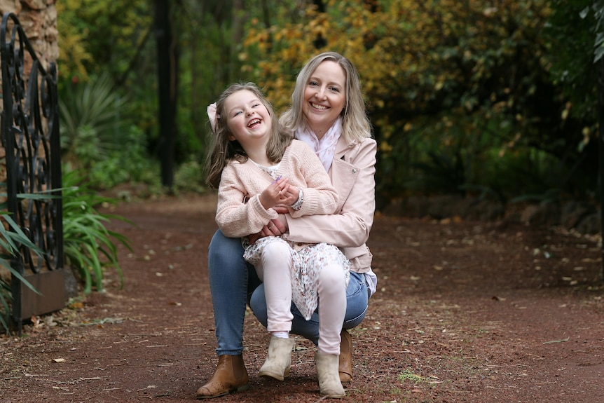 A blonde woman crouching with her daughter on her lap in a forest.