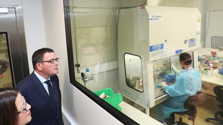 Premier Daniel Andrews peers through a glass wall at a scientist in researching a vaccine.