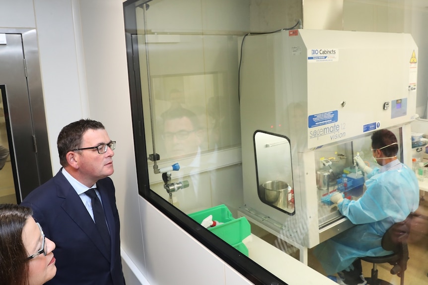 Premier Daniel Andrews peers through a glass wall at a scientist in researching a vaccine.