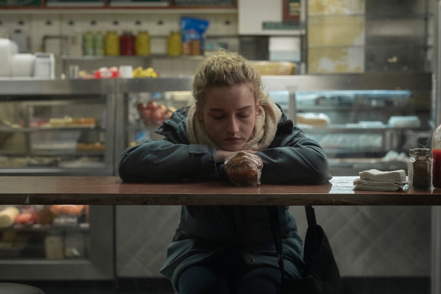 The actor Julia Garner in the film The Assistant, sitting at bar table in deli bent over looking at cling wrapped muffin.