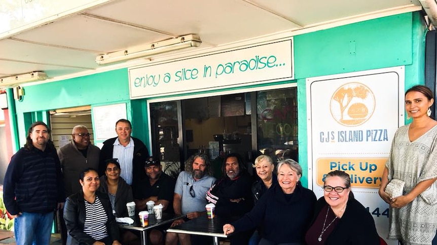 Twelve people smile at the camera, sitting and standing at a cafe with turquoise front walls