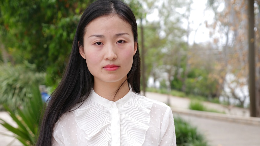 Ling Zhao looking at the camera, straight-faced. Wearing a white long-sleeved blouse, trees behind.