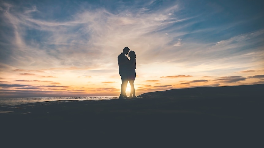 Silhouette of two people embracing on a clearing near the ocean with the sun on the horizon in a cloudy sky