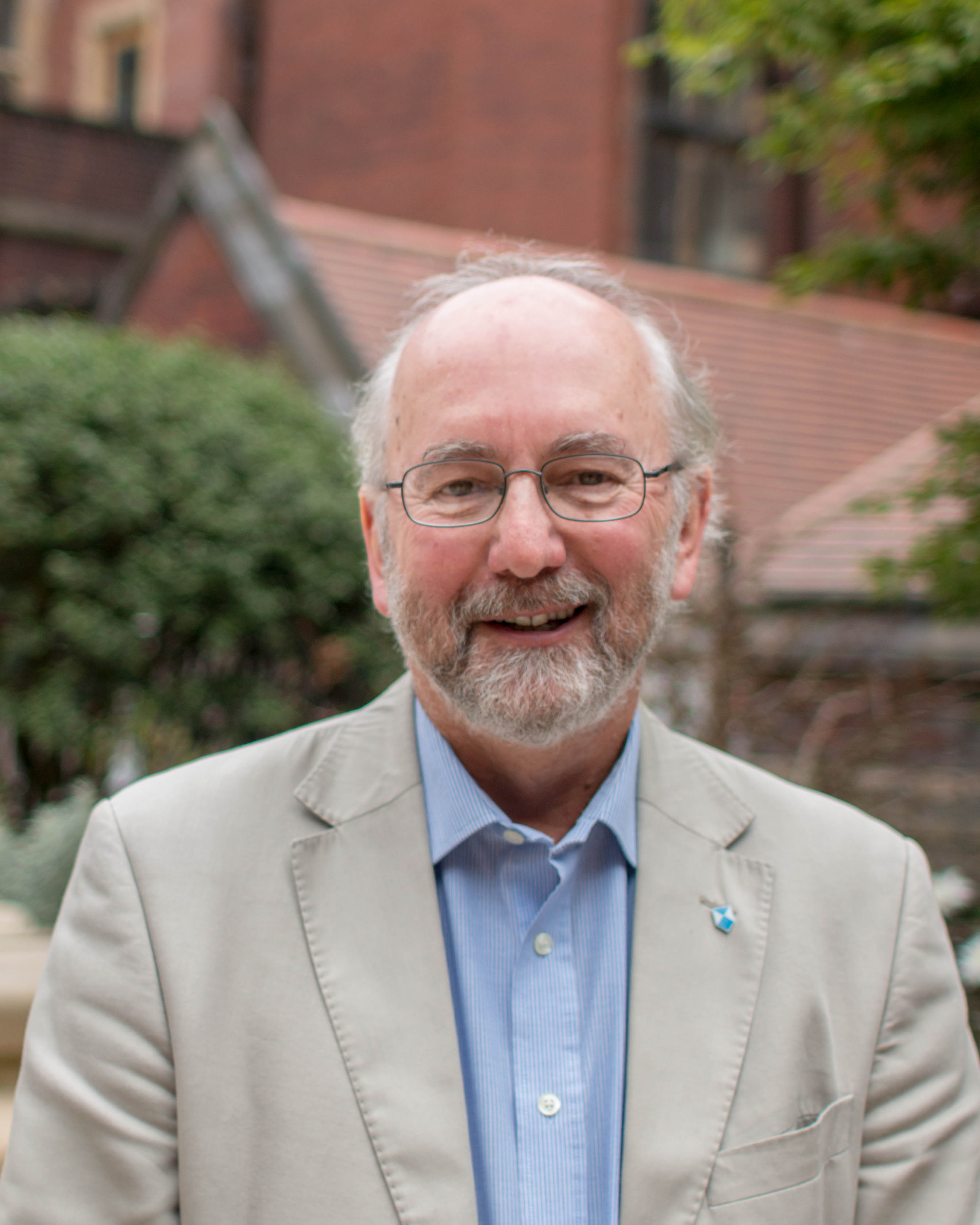 Peter Stone, an older man with a beard and glasses, dressed in a light jacket and button-down, smiles, standing outside.