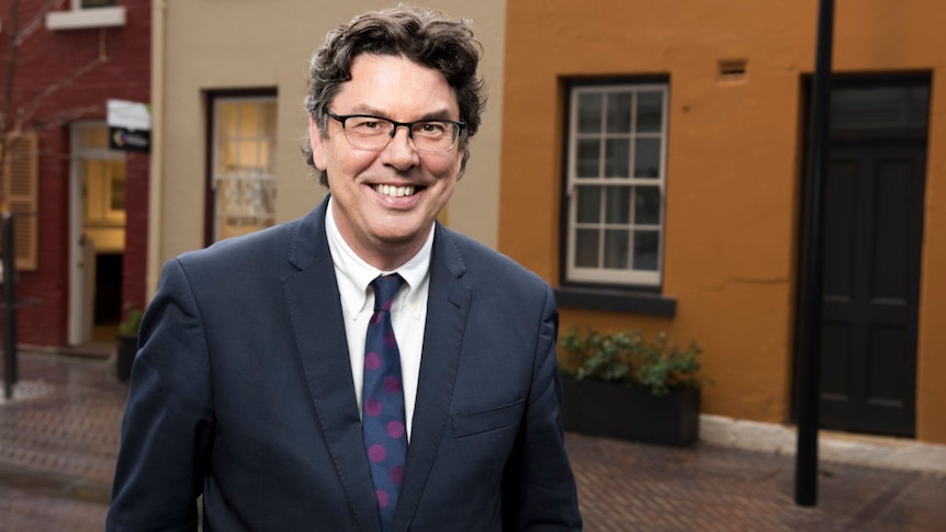 A man with brown, curly hair and glasses is smiling broadly. He's wearing a navy suit, white shirt and red-spotted navy tie