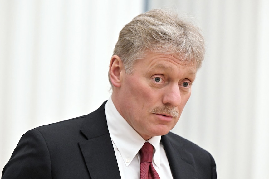 A 60-something man with sandy hair and moustache looks thoughtful in a dark suit with white shirt and red tie