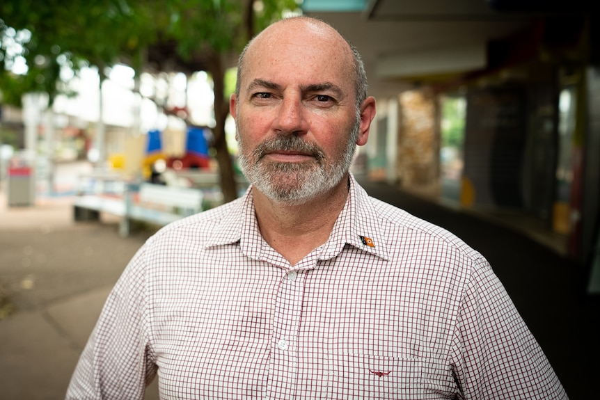 A man with a short beard wearing a button-down shirt looks seriously at the camera. he is outside in the Darwin CBD.