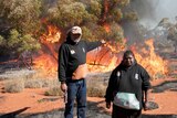 Two Indigenous people stand in the foreground as fire burns in scrub behind them.