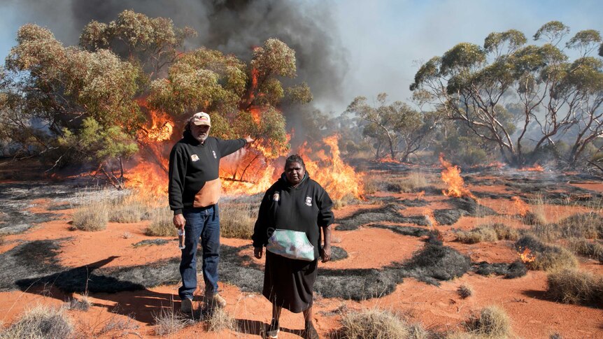 Two Indigenous people stand in the foreground as fire burns in scrub behind them.