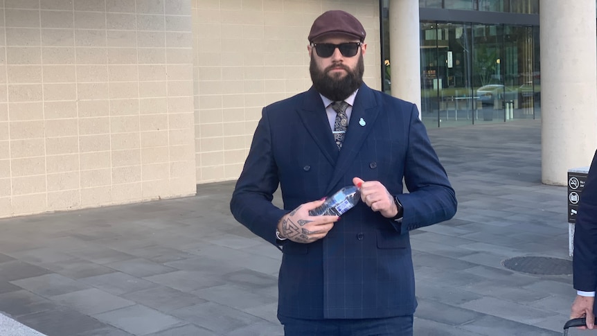 A man with a long groomed beard and cap, tattooed hands, wearing suit and sunglasses, walks out of a courthouse.