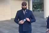 A man with a long groomed beard and cap, tattooed hands, wearing suit and sunglasses, walks out of a courthouse.
