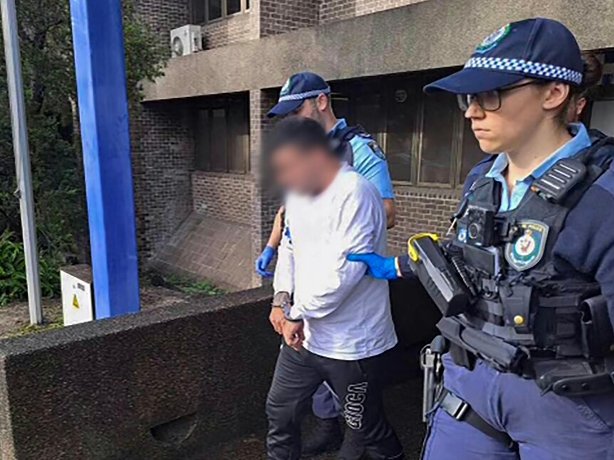 A man in a white long-sleeved t-shirt is escorted by two police officers while in handcuffs outside a police station.