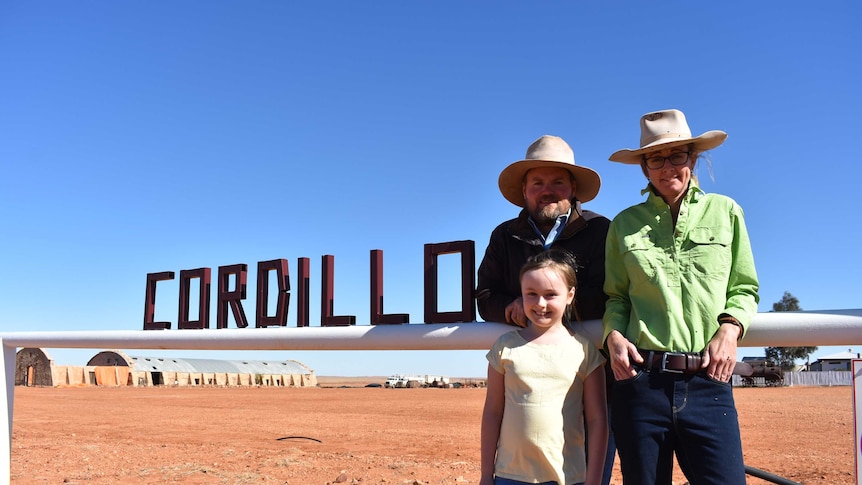 Two parents wearing broad hats stand next to a sign which says 'Cordillo' with their daughter in front of them.