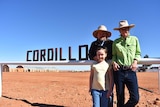 Two parents wearing broad hats stand next to a sign which says 'Cordillo' with their daughter in front of them.