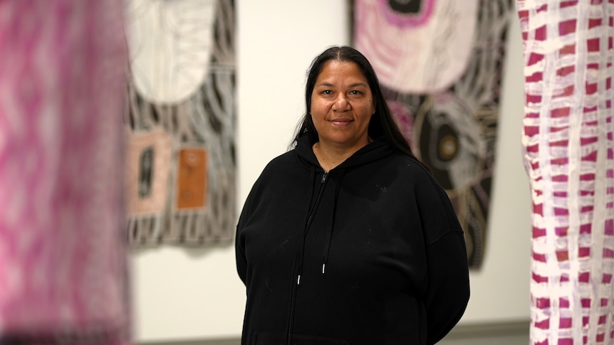 Rosalee is smiling wearing a blak zip up jumper. Behind her is two bark paintings with earthy tones and bright pink paint