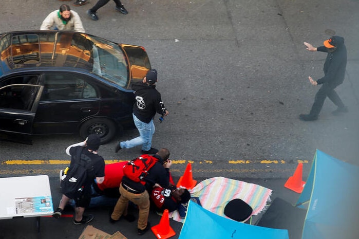 A man is seen walking around the back of a black car as medical workers tend to a shooting victim on the ground next to it.
