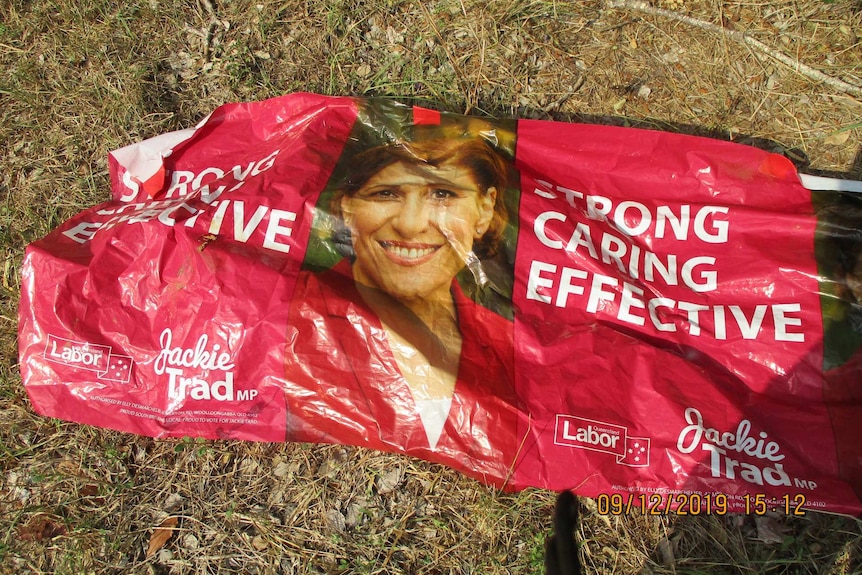 A red campaign sign with Jackie Trad's face on it lies scrunched and wrinkled on the grass