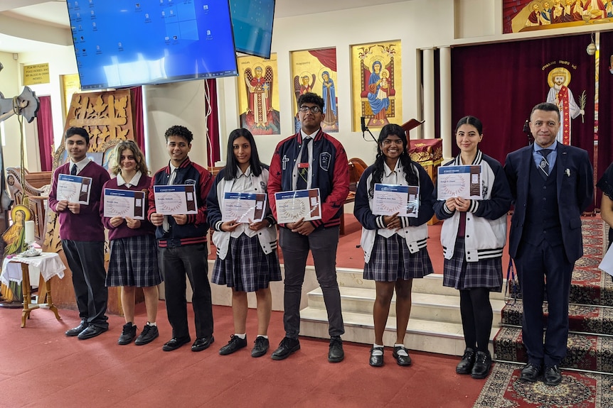 A long of uniformed students hold up paper awards. A suited man and woman stand on the right.