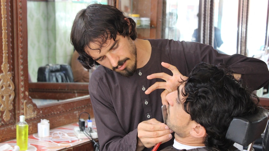 A barber wearing traditional Afghan attire cuts a man's beard in his shop