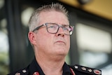 A man with a serious expression wearing black glasses and a navy NT Police uniform looks off camera.